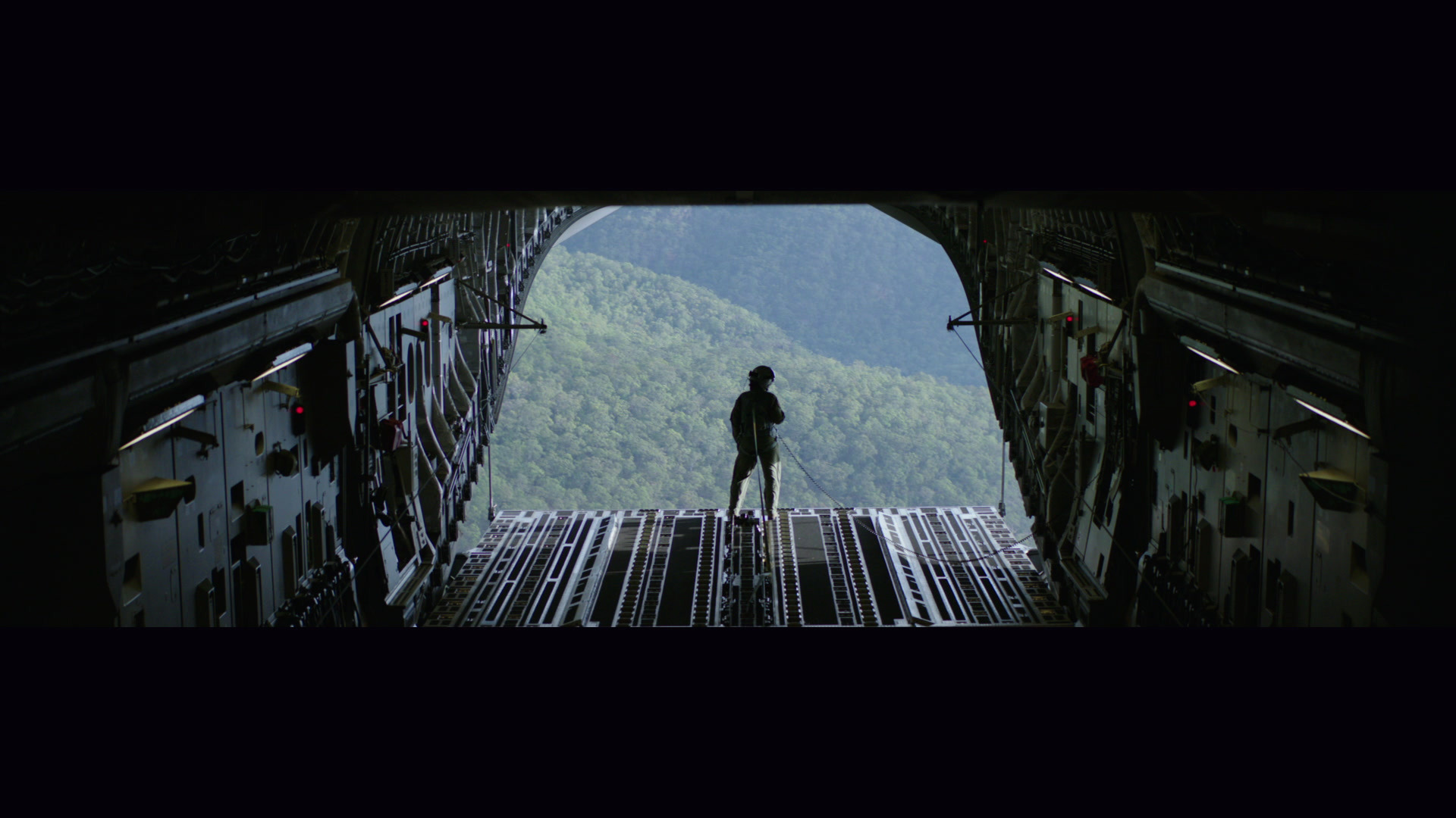 An airman stands on the open ramp of a transport aircraft above a forested ridge.